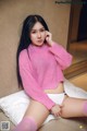 A woman in a pink sweater and white panties sitting on a bed.